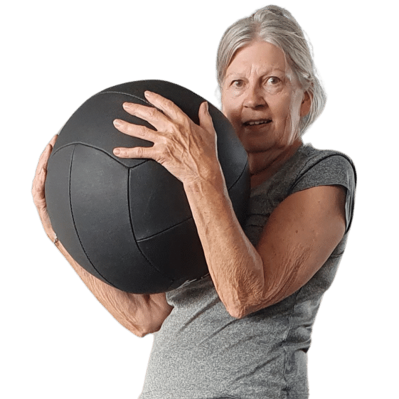 An older woman holding a large exercise ball, dressed in workout clothes, demonstrating strength and balance during a fitness session at Right Time PT in Leicester.